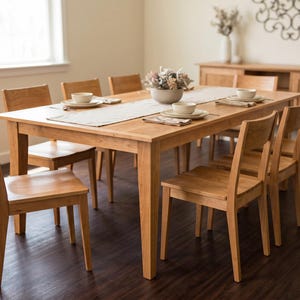 May include: A rectangular light brown wooden dining table with eight matching chairs. The table is set with a long white runner, white ceramic bowls on plates, and folded napkins. A central bowl contains a decorative floral arrangement.