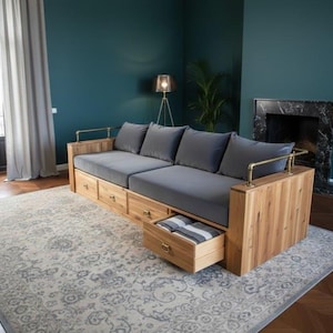 May include: A wooden sofa with gray cushions and built-in drawers. The sofa has a light wood frame with brass accents on the arms. The drawers are open, revealing folded blankets. The sofa sits on a patterned rug.