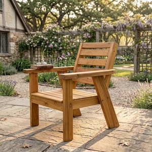 以下が含まれることがあります： 背もたれとアームレストが付いた木製の屋外用アームチェア。椅子は明るい色の木材でできており、石畳のパティオに置かれています。本と眼鏡がアームレストに置かれています。背景には庭と石造りの建物があります。