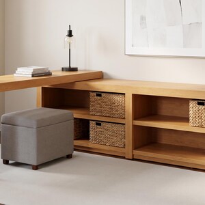 May include: A light wood desk with an attached bookcase and storage baskets. A gray upholstered ottoman sits in front of the desk. A lamp and books are on the desk. A white rug is on the floor.