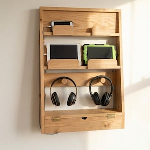 May include: A wall-mounted light wood organizer featuring two shelves with dividers for tablets and phones, two pegs for hanging headphones, and a bottom storage compartment with a hinged lid and brass-colored metal hinges.