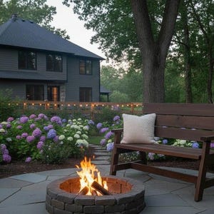 May include: Outdoor scene with a fire pit, wooden bench, and blooming hydrangeas. A house is in the background. The fire pit is made of brick and has a fire burning inside. The bench has a cream-colored pillow.