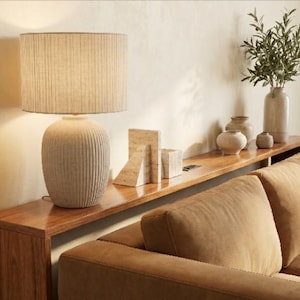 Op de afbeelding: Een tafellamp met een gestructureerde, geribbelde, gebroken witte keramische voet en een cilindrische stoffen lampenkap. De lamp verspreidt een warme gloed en werpt licht op de muur erachter. De voet heeft overal verticale groeven.