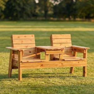 May include: A two-seater wooden bench with a small central table, situated on a grassy lawn. The bench is constructed from light-coloured wood with a natural finish. The background shows a green field and trees.