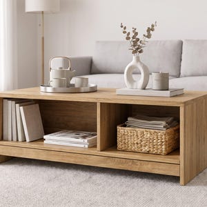 May include: A rectangular light oak coffee table featuring two open storage compartments. The left side holds several upright books and stacked magazines, while the right side contains a woven wicker basket filled with folded light-colored textiles.
