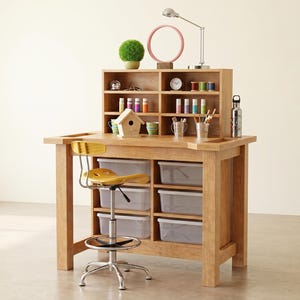 May include: A wooden craft desk with a yellow chair, storage bins, and a built-in shelf. The shelf holds colorful spools of thread, paint bottles, and a small birdhouse. A desk lamp and a pink circular mirror are on top.