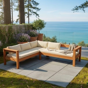 May include: An outdoor sectional sofa with a wooden frame and beige cushions. The L-shaped seating arrangement is on a gray stone patio, with a view of the ocean and trees in the background. A blanket and a book are on the sofa.