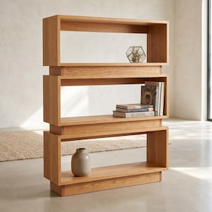 May include: A light brown wooden bookshelf featuring three open rectangular tiers stacked vertically. The top shelf holds a geometric glass terrarium, the middle shelf contains a stack of books, and the bottom shelf displays a single beige ceramic vase.