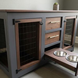 Puede incluir: Una jaula para perros de madera con marco gris y detalles marrones. Cuenta con puertas de malla metálica, dos cajones con tiradores dorados y una bandeja extraíble con dos cuencos de acero inoxidable para comida y agua. Una pequeña planta encima.