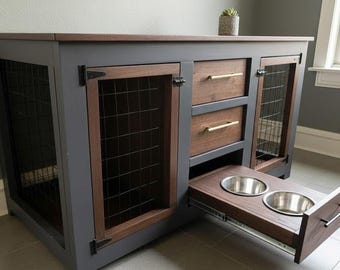 May include: A wooden dog crate with a gray frame and brown accents. It features wire mesh doors, two drawers with gold handles, and a pull-out tray with two stainless steel bowls for food and water. A small plant sits on top.