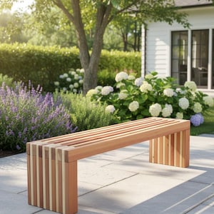 May include: A wooden bench with a slatted seat and vertical support beams. The bench is made of light brown wood and is set on a stone patio. The background features a lush garden with green foliage and flowering plants.