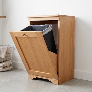 Puede incluir: Un mueble basculante para basura hecho de madera de tono claro con un marco rectangular y un panel frontal único que se abre hacia afuera sobre bisagras para revelar un contenedor de basura de plástico negro en su interior con un pomo redondo.