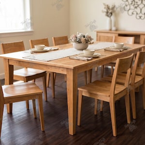 May include: A rectangular light brown wooden dining table with six matching chairs. The table is set with a white runner, white ceramic bowls on plates, and folded napkins. The furniture features a smooth finish and straight legs.