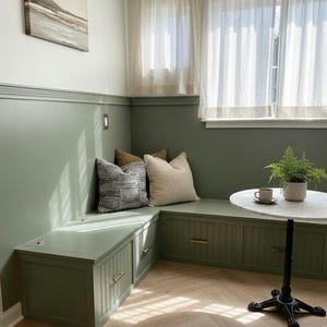 May include: A built-in bench with storage, painted in a muted green, features three decorative pillows. A small round table with a marble top and a fern in a pot sit nearby. The room has a cozy, inviting atmosphere.