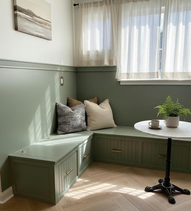 May include: A built-in bench with storage, painted in a muted green, features three decorative pillows. A small round table with a marble top and a fern in a pot sit nearby. The room has a cozy, inviting atmosphere.