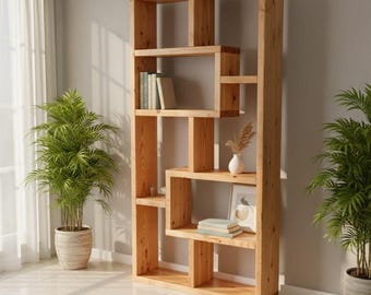 Puede incluir: Una estantería alta de madera clara con un diseño asimétrico. Los estantes contienen libros, un pequeño jarrón blanco con flores secas y una imagen enmarcada. Dos plantas en macetas flanquean la estantería, añadiendo un toque de vegetación.