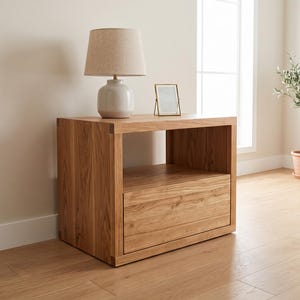 Peut inclure: Une table de chevet rectangulaire en bois avec une finition naturelle en grain de bois brun clair, une étagère supérieure ouverte et un tiroir inférieur. Une lampe avec un abat-jour en tissu beige et une base en céramique claire est posée dessus.