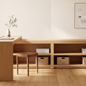 May include: A light-colored wooden desk with built-in shelving and a matching stool. The desk has a raised section with a small vase and a laptop. Woven baskets and white boxes are placed on the shelves. A framed artwork hangs on the wall.