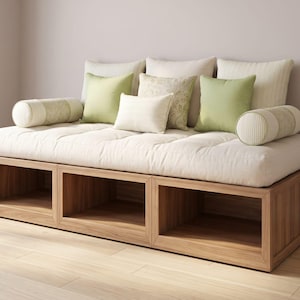 May include: A wooden daybed with a cream-colored mattress and several pillows in shades of green and cream. The base of the daybed has three open storage compartments. The room has light-colored walls and a wooden floor.