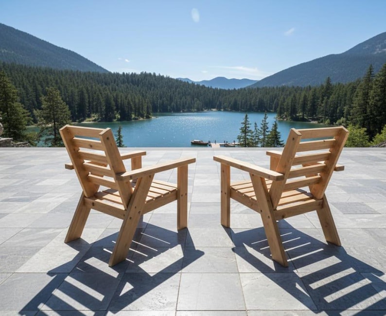 以下が含まれることがあります： アームレスト付きの木製椅子が2脚、石畳のパティオに置かれ、湖と山脈を望んでいます。椅子は明るい色の木材で作られており、日光の下で影を落としています。