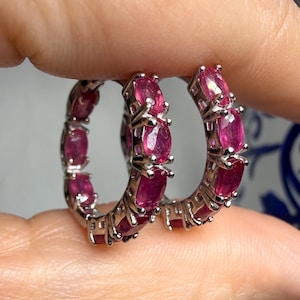 May include: A pair of hoop earrings featuring oval-cut, vibrant red gemstones set in a silver metal. The earrings are held up, showcasing the gemstones' color and the intricate setting. The design is elegant and eye-catching.