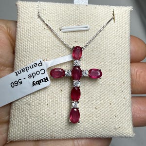May include: A silver necklace with a cross pendant. The cross is adorned with oval-shaped, deep red rubies and small, round, clear gemstones. The pendant hangs on a delicate silver chain. A tag reads "Ruby Pendant Code - 560".