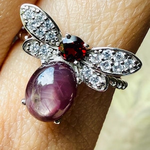 May include: A silver-toned ring shaped like a bee, featuring a large oval cabochon-cut deep purple stone for the body, a round faceted dark red stone for the head, and clear white stones embedded in the wings for a sparkling effect.