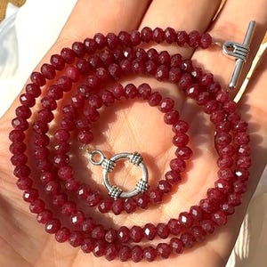 May include: A necklace composed of small, faceted, deep red beads arranged in a circular pattern. The piece features a silver-toned toggle clasp with a circular ring and a T-bar component, providing a secure closure for the beaded strand.