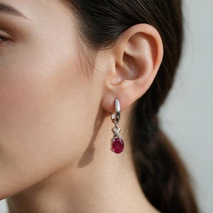 May include: A silver hoop earring featuring a dangling oval-cut deep red ruby gemstone. The setting above the ruby is accented with small clear stones arranged in a cross pattern, connecting the hoop to the central red stone.