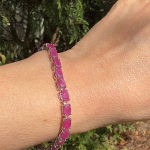 May include: A silver bracelet with oval-cut, vibrant pink gemstones. The bracelet is worn on a wrist, showcasing the gemstones' color and the intricate setting. The background is blurred, with green foliage.