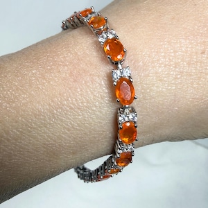 May include: A silver bracelet featuring oval-cut orange gemstones and small white accent stones. The bracelet is a tennis style, with the orange stones set in a row and separated by the white stones.