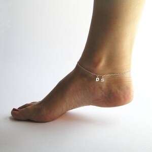 May include: A silver ankle chain with two dangling charms, the letters "D" and "S", on a person's ankle.
