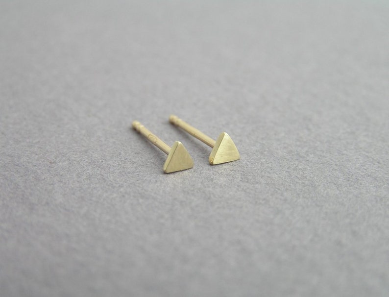 May include: A pair of gold triangle stud earrings. The earrings are small and delicate, and they have a simple, minimalist design.
