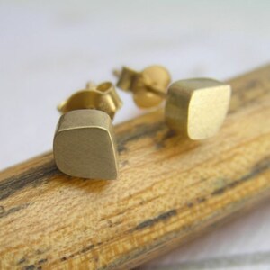 May include: A pair of gold-toned square stud earrings. The earrings are made of a smooth, brushed metal and have a slightly rounded edge.