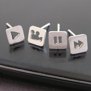 Op de afbeelding: Vier zilveren oorbellen met verschillende mediasymbolen: afspelen, videocamera, pauzeren en snel vooruit.