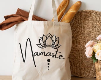 May include: Cream-colored tote bag with the word "Namaste" in a script font, accompanied by a black lotus flower design. The bag contains a brown cloth, two baguettes, and is next to a woven mat and a basket of flowers.