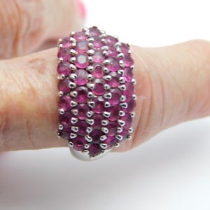 May include: A silver ring featuring a wide, multi-row band densely set with numerous small, round, deep pinkish-red faceted gemstones. The stones are arranged in vertical columns and secured with small metal prongs across the surface.