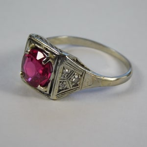 May include: A vintage ring featuring a bright magenta gemstone in a rectangular, ornate, white metal setting. The band has intricate detailing and small accent stones. A classic and elegant piece of jewellery. The ring is a piece of jewellery.