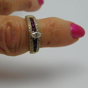 May include: A gold-toned ring featuring a central marquise-cut diamond, flanked by rows of small, vibrant red gemstones and clear accent stones. The ring is set on a finger with pink nail polish.