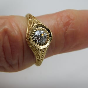May include: A gold filigree ring with a central, round, clear gemstone. The ring is set on a finger with a bright pink nail polish. The ring has an ornate, detailed design.