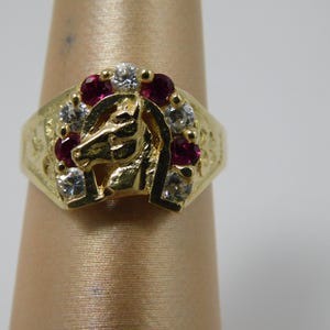 Puede incluir: Un anillo dorado con un diseño de cabeza de caballo. El anillo está adornado con piedras preciosas claras y rojas. La cabeza del caballo está centrada dentro de una herradura, con piedras preciosas que rodean el diseño.