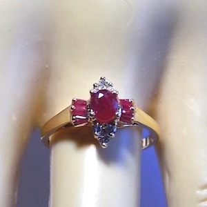 Puede incluir: Un anillo de oro con un rubí central ovalado, flanqueado por dos rubíes más pequeños y acentuado con pequeños diamantes. El anillo tiene una banda de oro, mostrando un diseño clásico y elegante. Las vibrantes piedras preciosas crean un contraste llamativo.