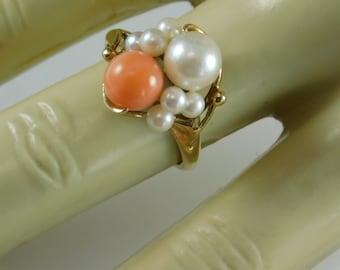 Può includere: Un anello d'oro con un gruppo di perle e una perla di corallo. L'anello ha un design unico con una grande perla bianca, una perla di corallo e diverse perle più piccole. L'anello è montato su una fascia d'oro.
