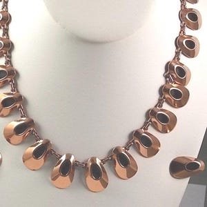 May include: A copper-toned necklace and matching earrings set. The necklace features a series of teardrop-shaped links with a dark oval inset. The earrings mirror the necklace design. The jewelry is displayed on a white mannequin bust.