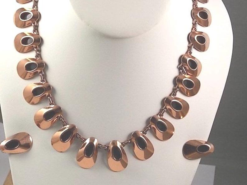 May include: A copper-toned necklace and matching earrings set. The necklace features a series of teardrop-shaped links with a dark oval inset. The earrings mirror the necklace design. The jewelry is displayed on a white mannequin bust.