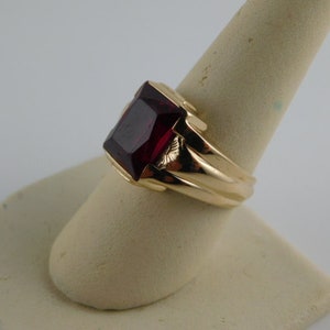 May include: A gold ring featuring a large, rectangular, deep red gemstone. The ring has a thick band and a detailed setting that holds the stone. The ring is displayed on a neutral-colored ring holder.