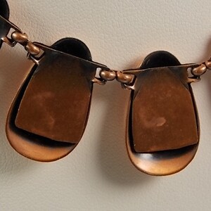 May include: A copper-toned necklace with a series of teardrop-shaped pendants. Each pendant has a layered design, with a darker, almost black, interior. The pendants are connected by small, round links.