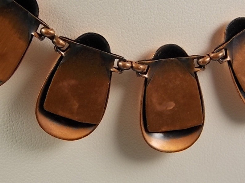 May include: A copper-toned necklace with a series of teardrop-shaped pendants. Each pendant has a layered design, with a darker, almost black, interior. The pendants are connected by small, round links.