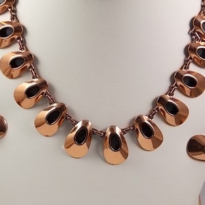 May include: A copper-toned necklace and matching earrings set. The necklace features a series of teardrop-shaped pendants with black oval insets, connected by small links. The earrings mirror the pendant design. The jewelry is displayed on a white bust.