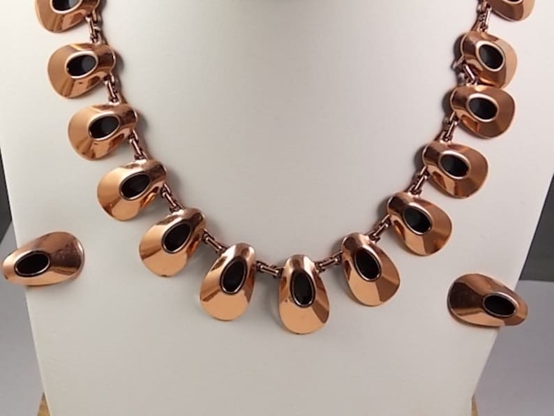May include: A copper-toned necklace and matching earrings set. The necklace features a series of teardrop-shaped pendants with black oval insets, connected by small links. The earrings mirror the pendant design. The jewelry is displayed on a white bust.